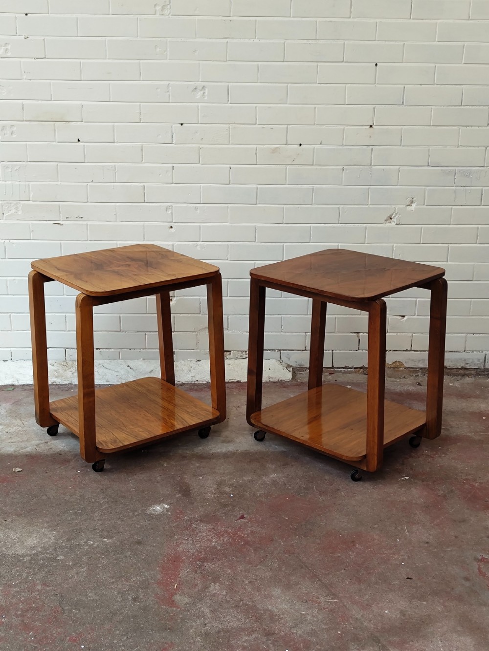 A matching pair of two-tier Art Deco burr walnut bedside tables featuring rounded tops, lower shelves, curved upright legs, and small castor wheels. View XI