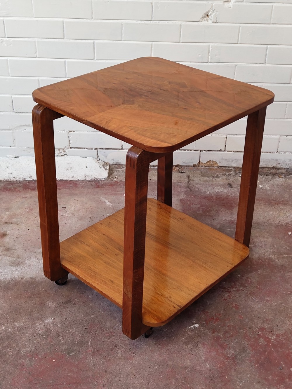 A matching pair of two-tier Art Deco burr walnut bedside tables featuring rounded tops, lower shelves, curved upright legs, and small castor wheels. View VI