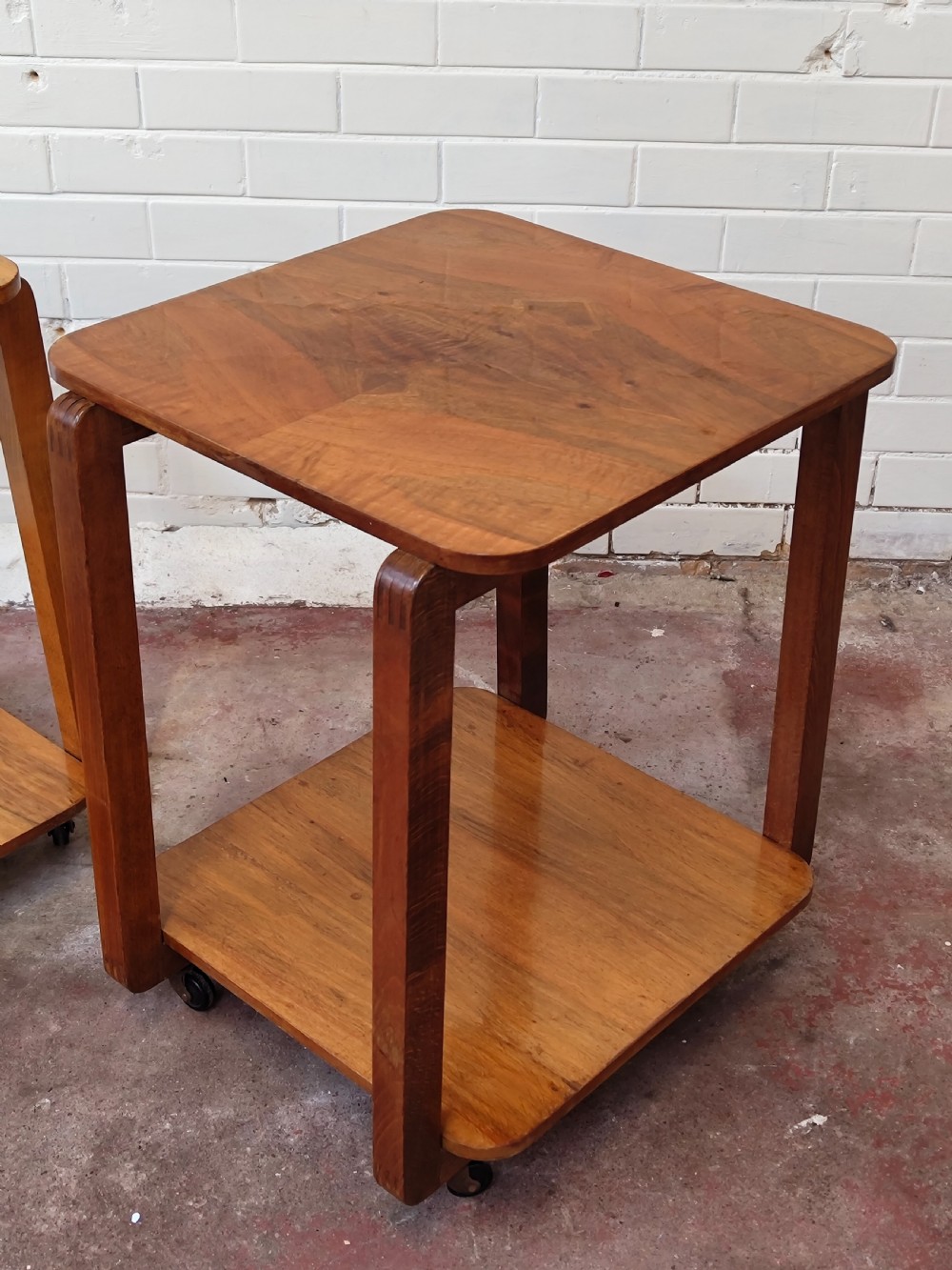A matching pair of two-tier Art Deco burr walnut bedside tables featuring rounded tops, lower shelves, curved upright legs, and small castor wheels. View VII