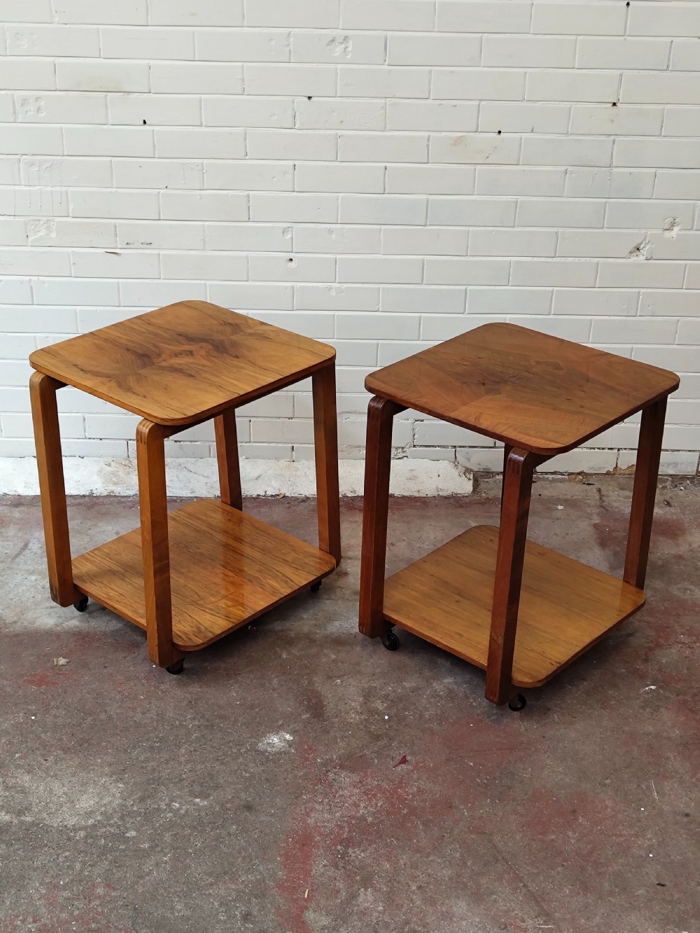 A matching pair of two-tier Art Deco burr walnut bedside tables featuring rounded tops, lower shelves, curved upright legs, and small castor wheels. View X