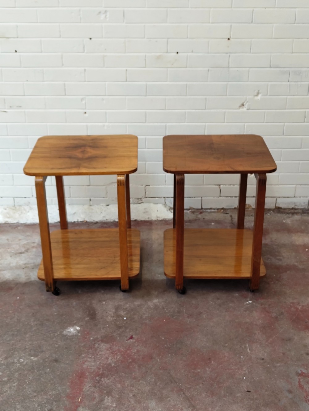 A matching pair of two-tier Art Deco burr walnut bedside tables featuring rounded tops, lower shelves, curved upright legs, and small castor wheels.