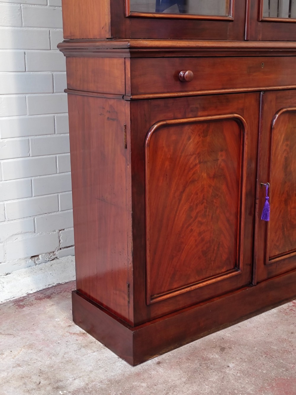 A tall Victorian mahogany bookcase featuring a glazed upper display cabinet with adjustable shelves over a lower two-door cupboard with flame mahogany veneer panels. View IV