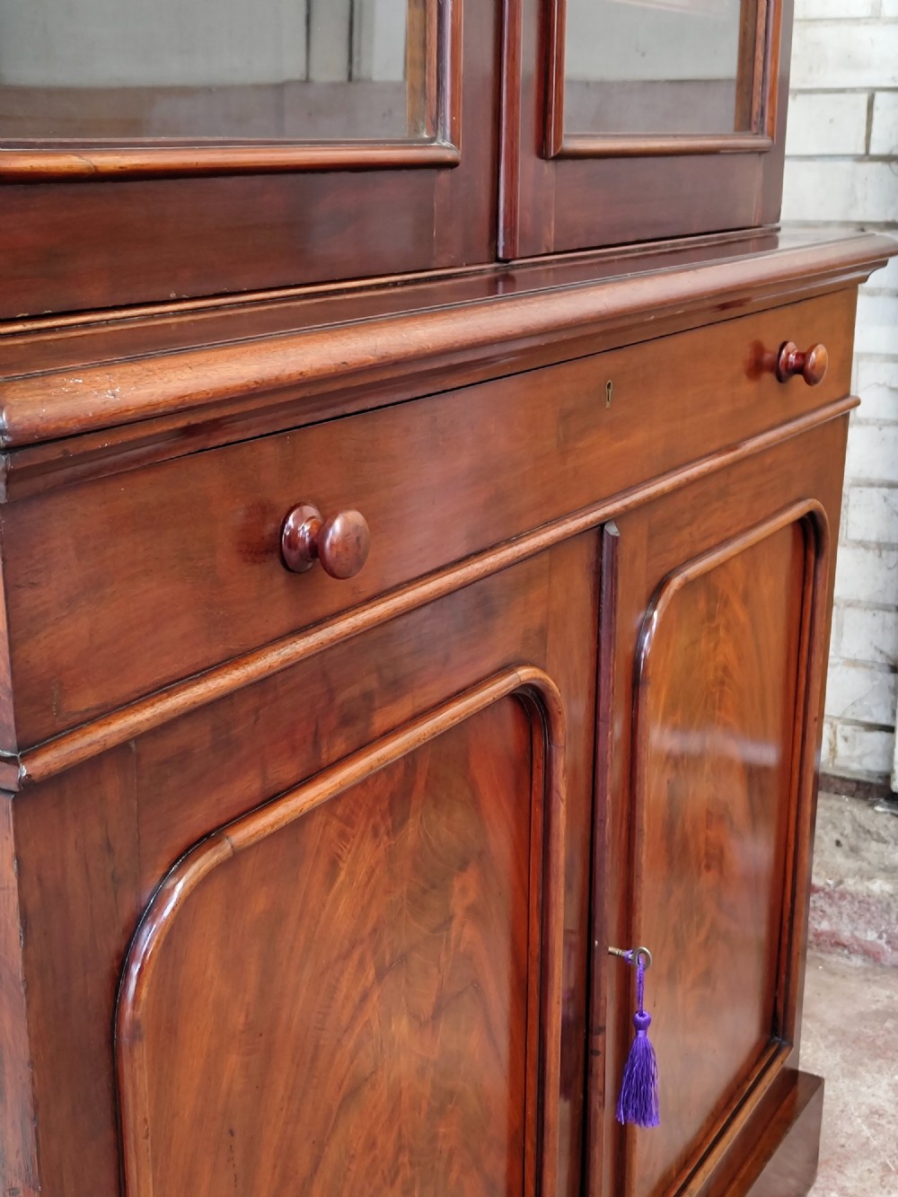 A tall Victorian mahogany bookcase featuring a glazed upper display cabinet with adjustable shelves over a lower two-door cupboard with flame mahogany veneer panels. View VI