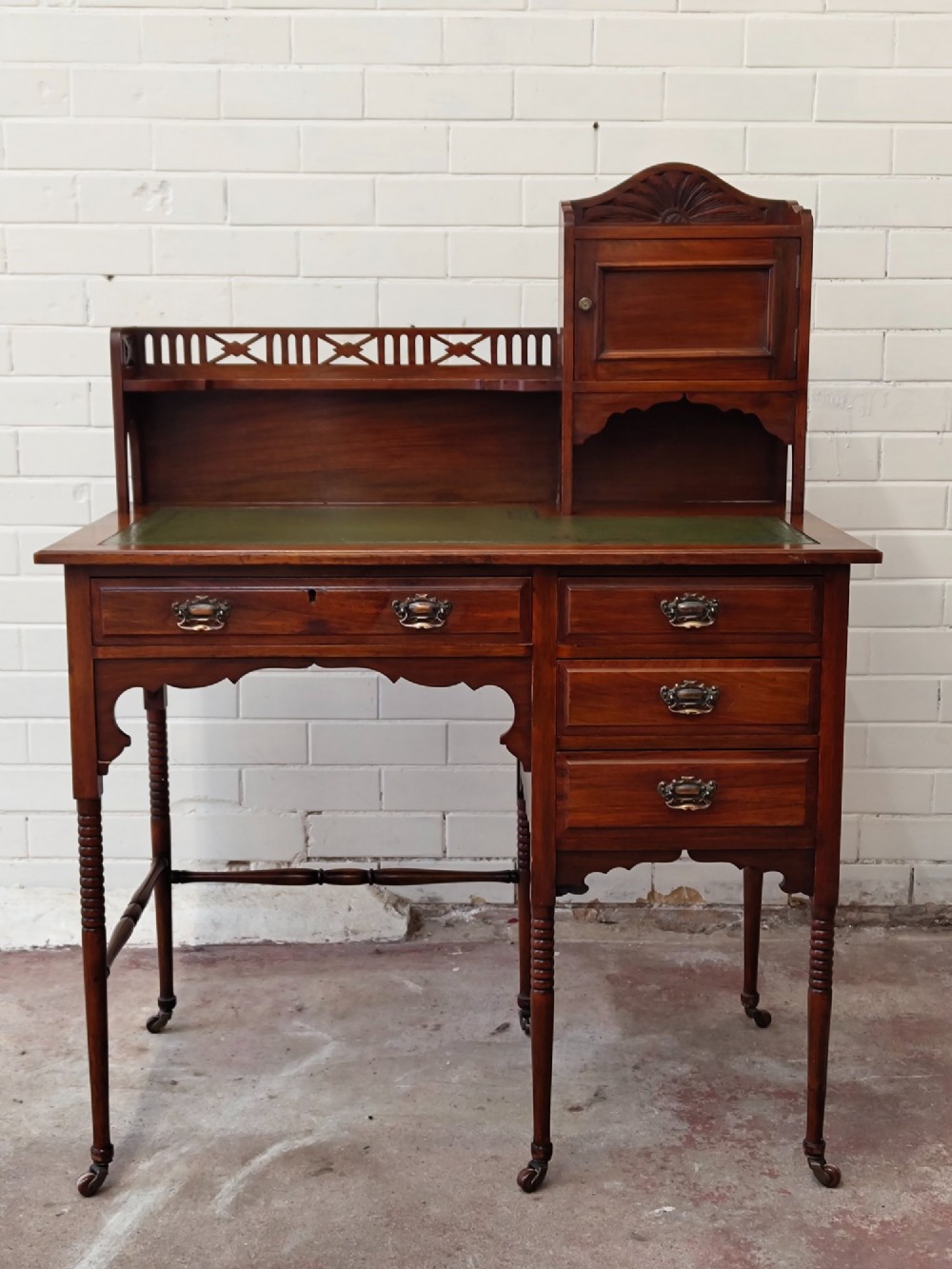An Edwardian mahogany ladies writing desk featuring a green leather writing surface, an asymmetric upper cabinet with gallery shelving, multiple drawers with brass handles, and slender turned legs on castors.