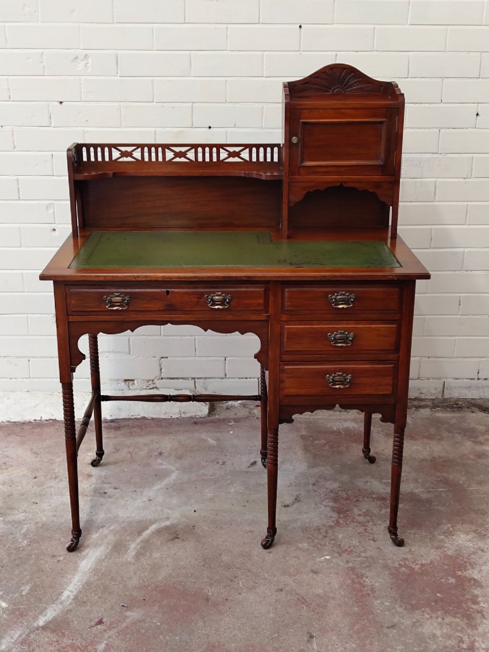 An Edwardian mahogany ladies writing desk featuring a green leather writing surface, an asymmetric upper cabinet with gallery shelving, multiple drawers with brass handles, and slender turned legs on castors. View II