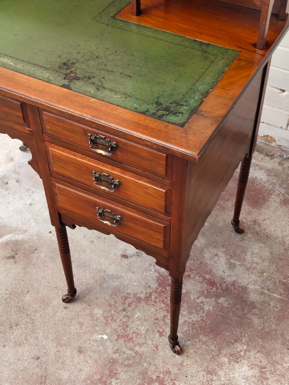 An Edwardian mahogany ladies writing desk featuring a green leather writing surface, an asymmetric upper cabinet with gallery shelving, multiple drawers with brass handles, and slender turned legs on castors. View IV
