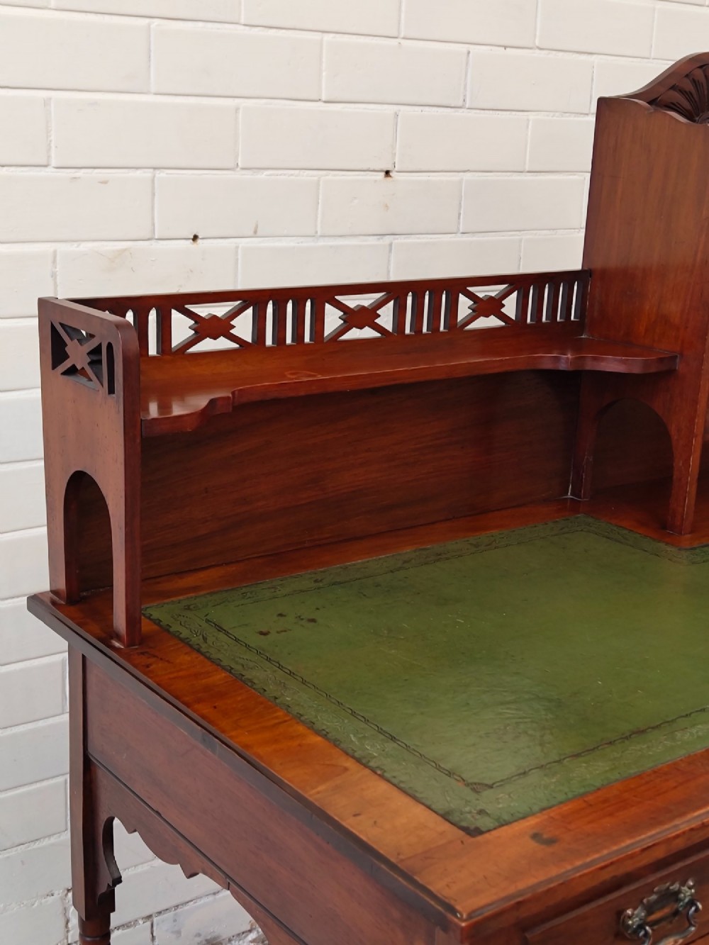 An Edwardian mahogany ladies writing desk featuring a green leather writing surface, an asymmetric upper cabinet with gallery shelving, multiple drawers with brass handles, and slender turned legs on castors. View VI