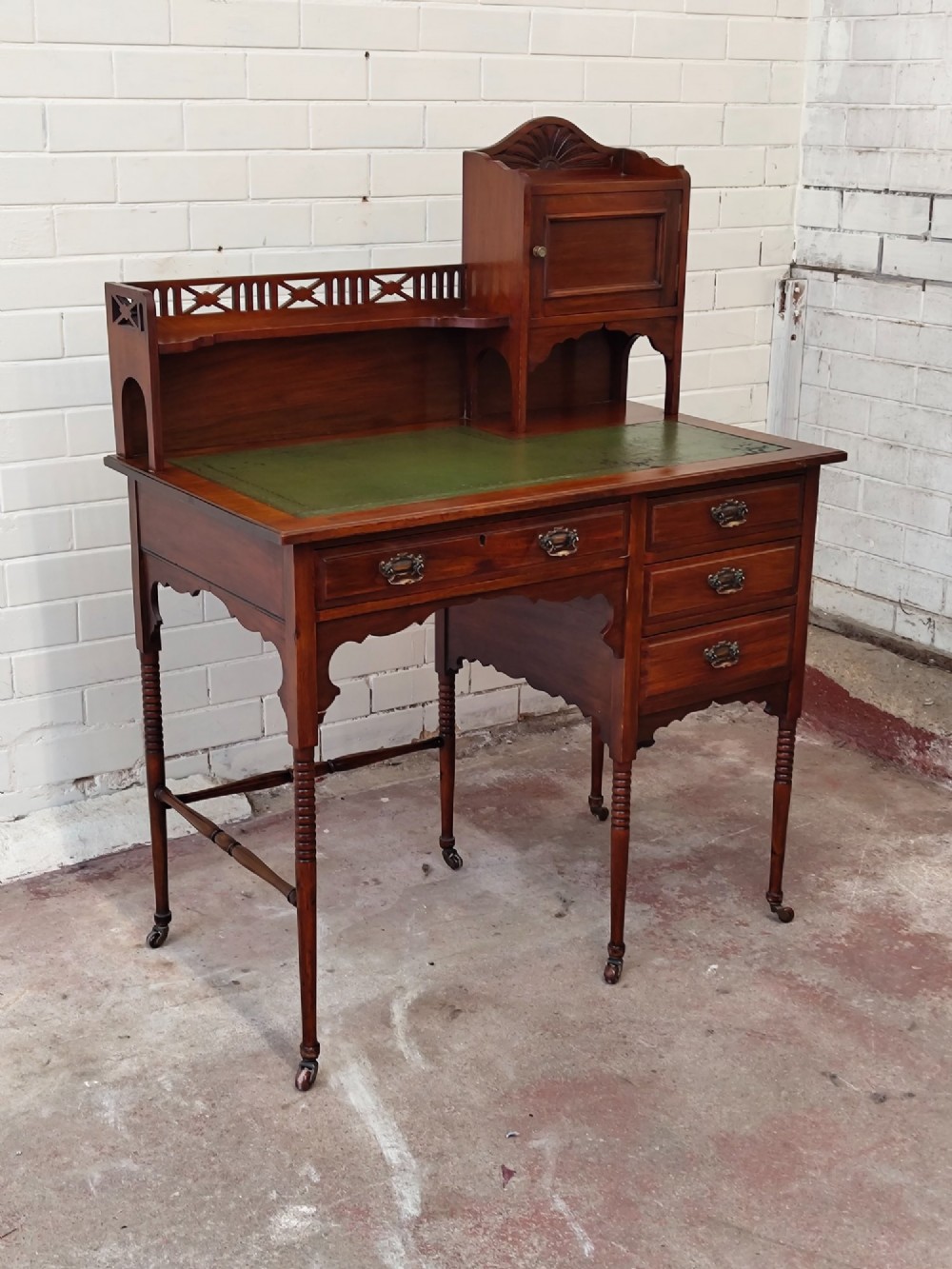An Edwardian mahogany ladies writing desk featuring a green leather writing surface, an asymmetric upper cabinet with gallery shelving, multiple drawers with brass handles, and slender turned legs on castors. View VII