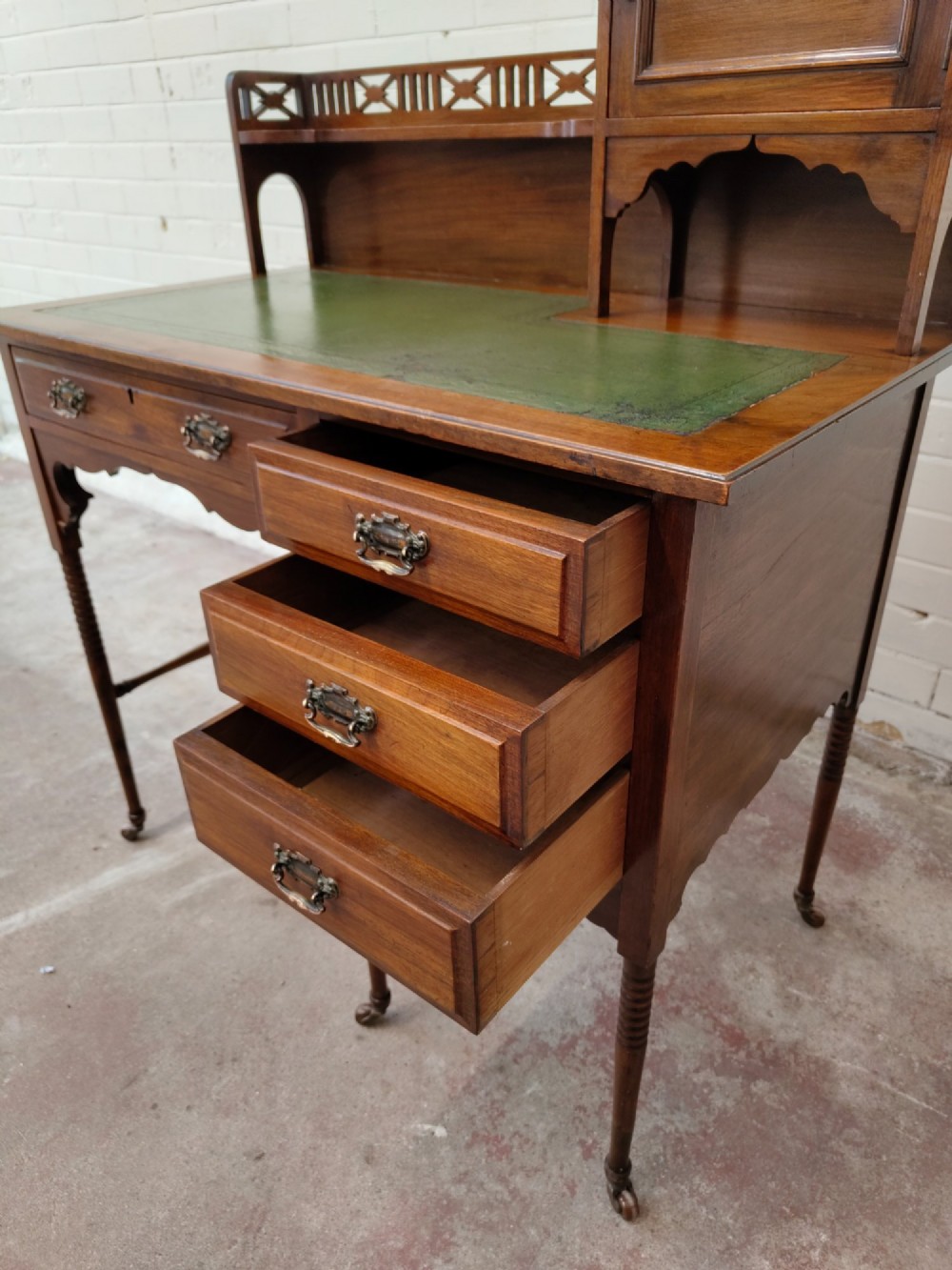 An Edwardian mahogany ladies writing desk featuring a green leather writing surface, an asymmetric upper cabinet with gallery shelving, multiple drawers with brass handles, and slender turned legs on castors. View VIII
