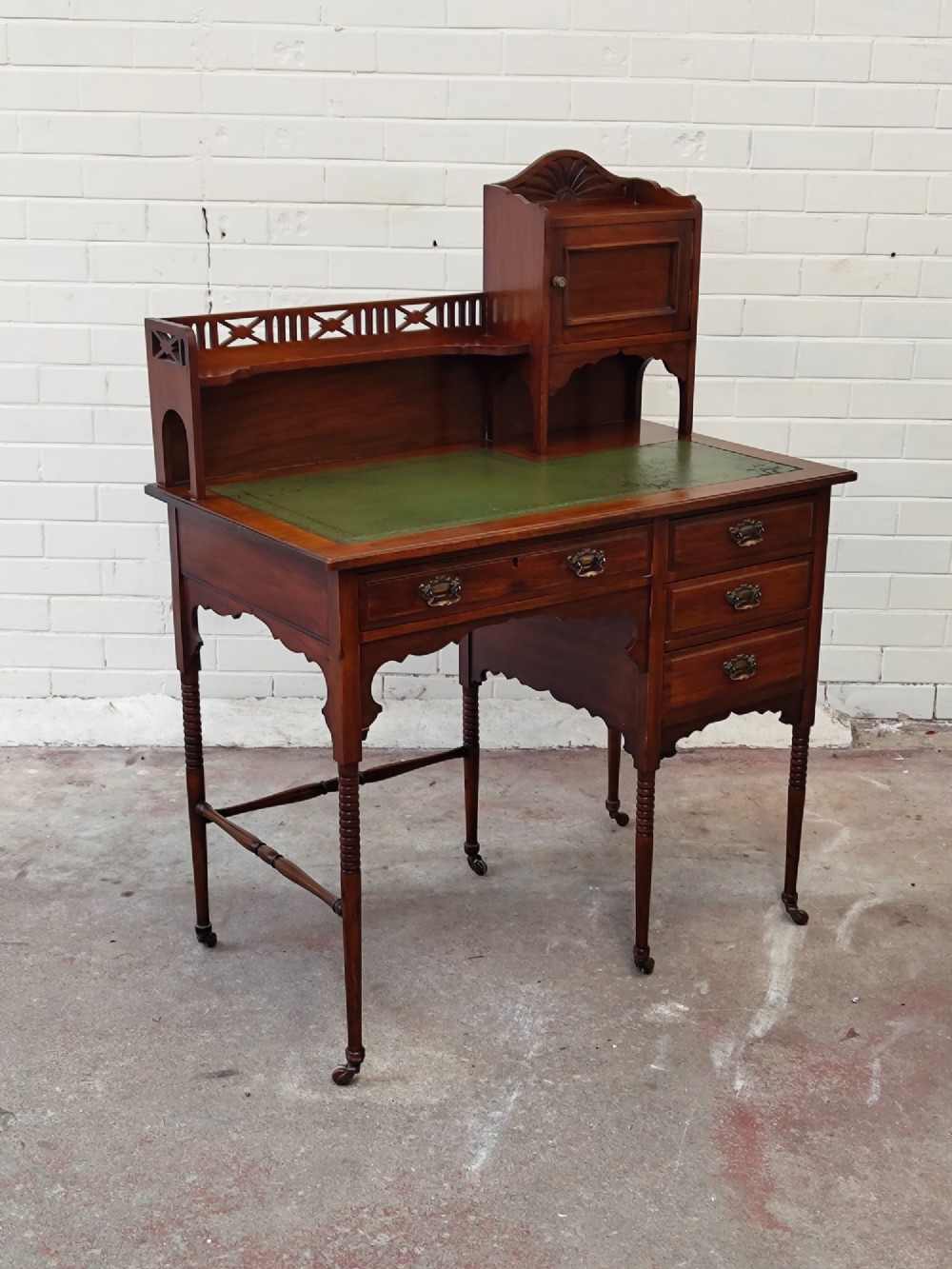 An Edwardian mahogany ladies writing desk featuring a green leather writing surface, an asymmetric upper cabinet with gallery shelving, multiple drawers with brass handles, and slender turned legs on castors. View IX