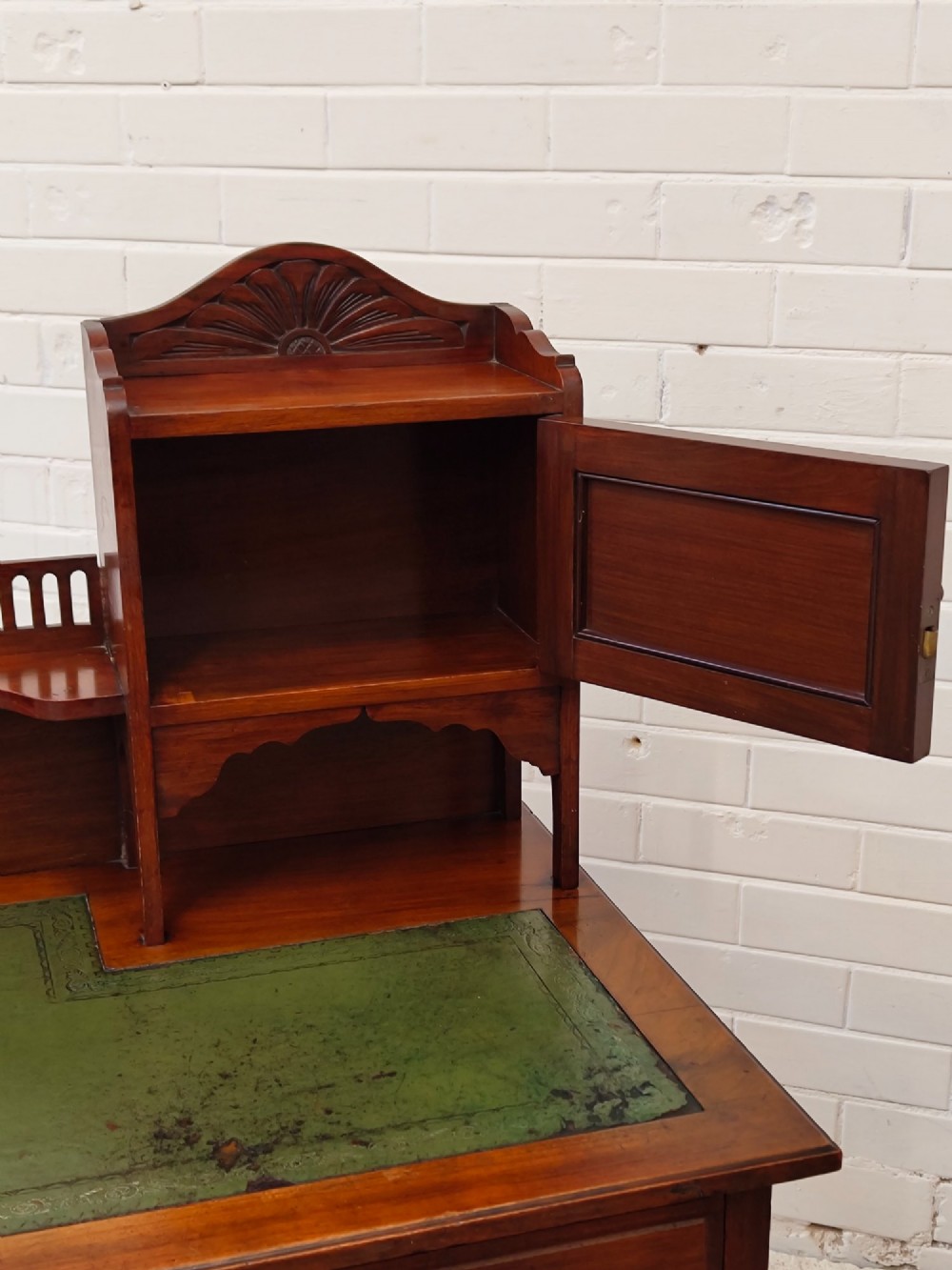 An Edwardian mahogany ladies writing desk featuring a green leather writing surface, an asymmetric upper cabinet with gallery shelving, multiple drawers with brass handles, and slender turned legs on castors. View X
