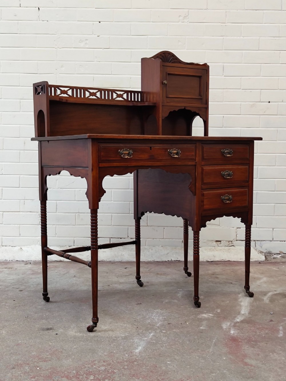 An Edwardian mahogany ladies writing desk featuring a green leather writing surface, an asymmetric upper cabinet with gallery shelving, multiple drawers with brass handles, and slender turned legs on castors. View XII