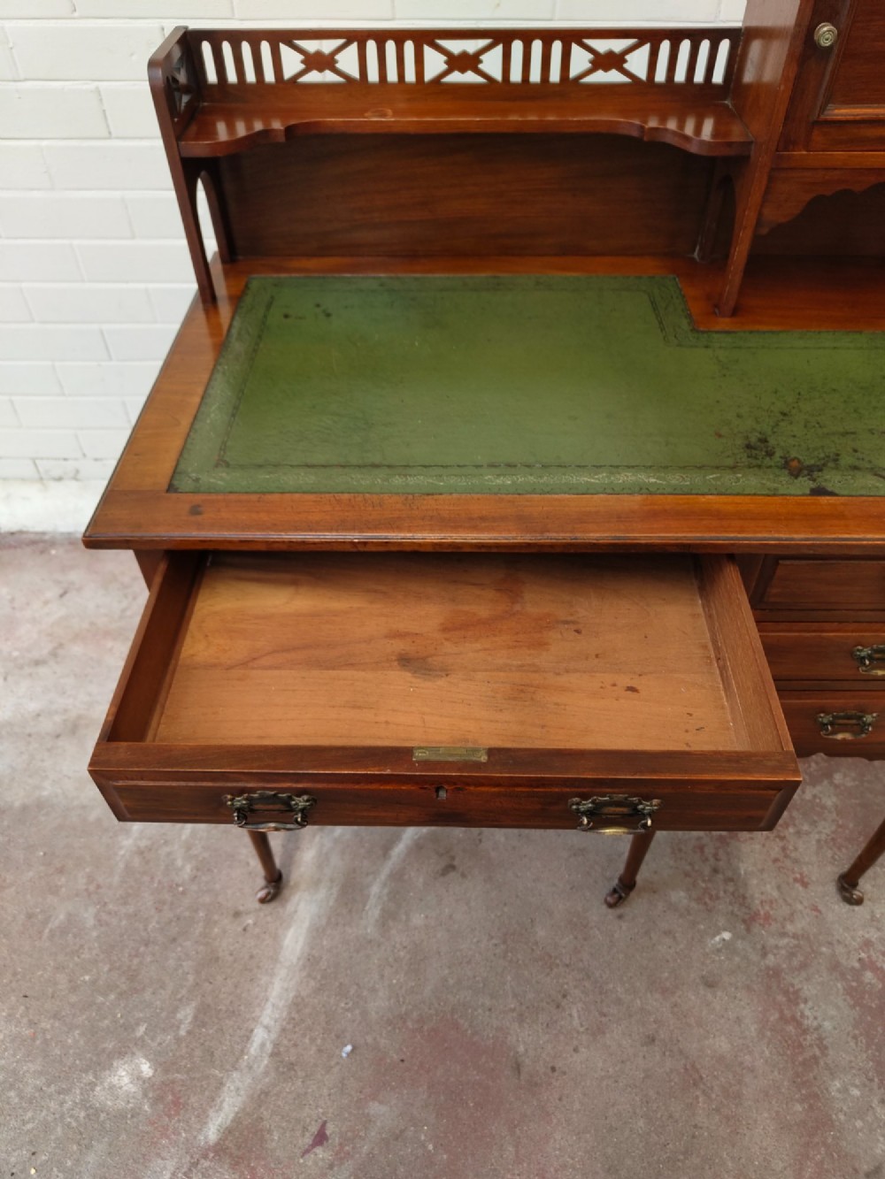 An Edwardian mahogany ladies writing desk featuring a green leather writing surface, an asymmetric upper cabinet with gallery shelving, multiple drawers with brass handles, and slender turned legs on castors. View XIII