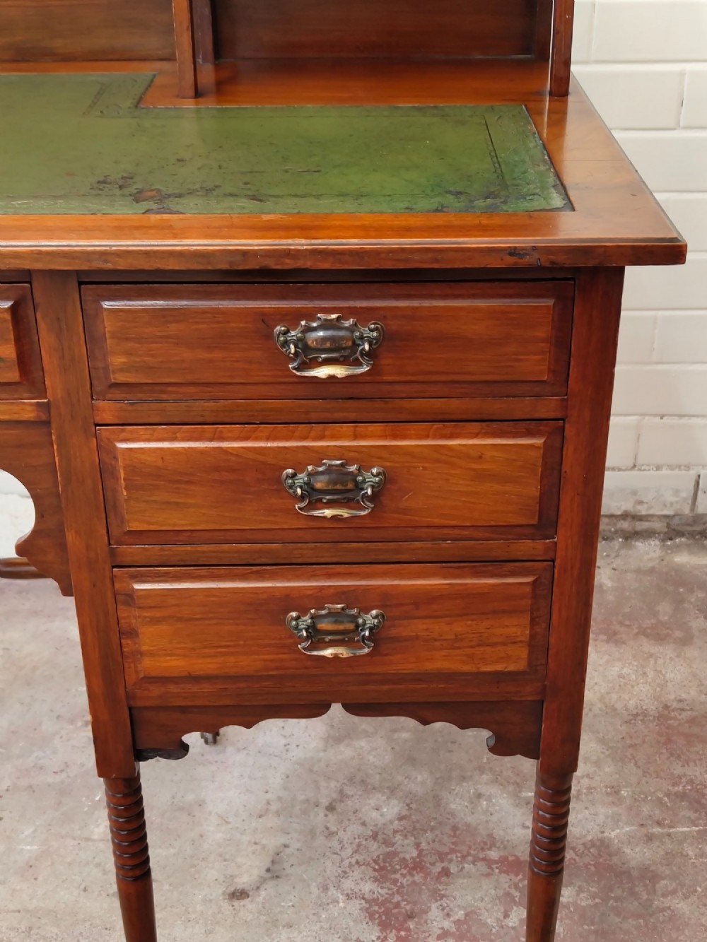 An Edwardian mahogany ladies writing desk featuring a green leather writing surface, an asymmetric upper cabinet with gallery shelving, multiple drawers with brass handles, and slender turned legs on castors. View XIV