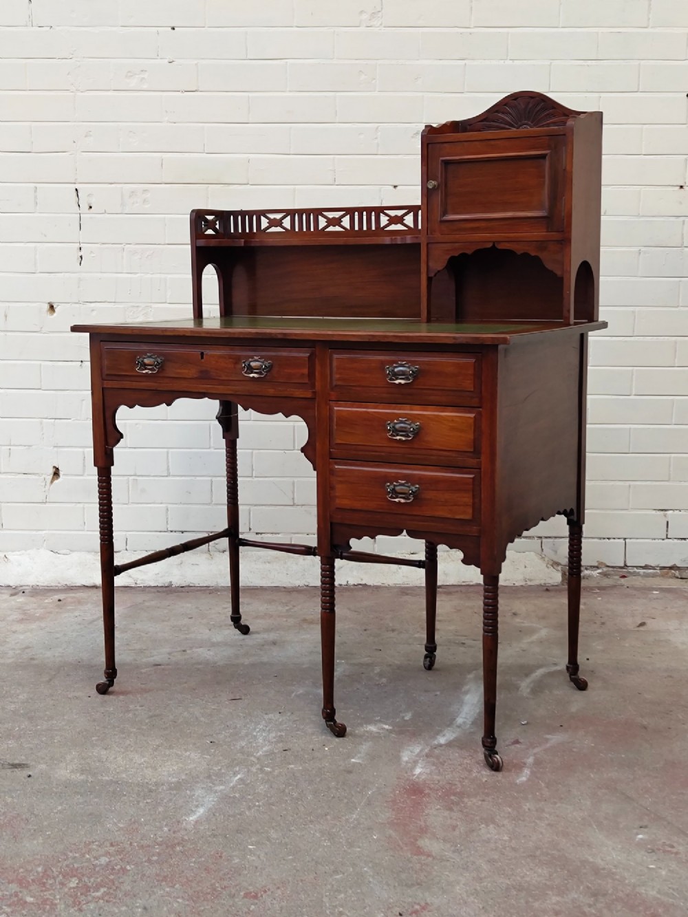 An Edwardian mahogany ladies writing desk featuring a green leather writing surface, an asymmetric upper cabinet with gallery shelving, multiple drawers with brass handles, and slender turned legs on castors. View XV