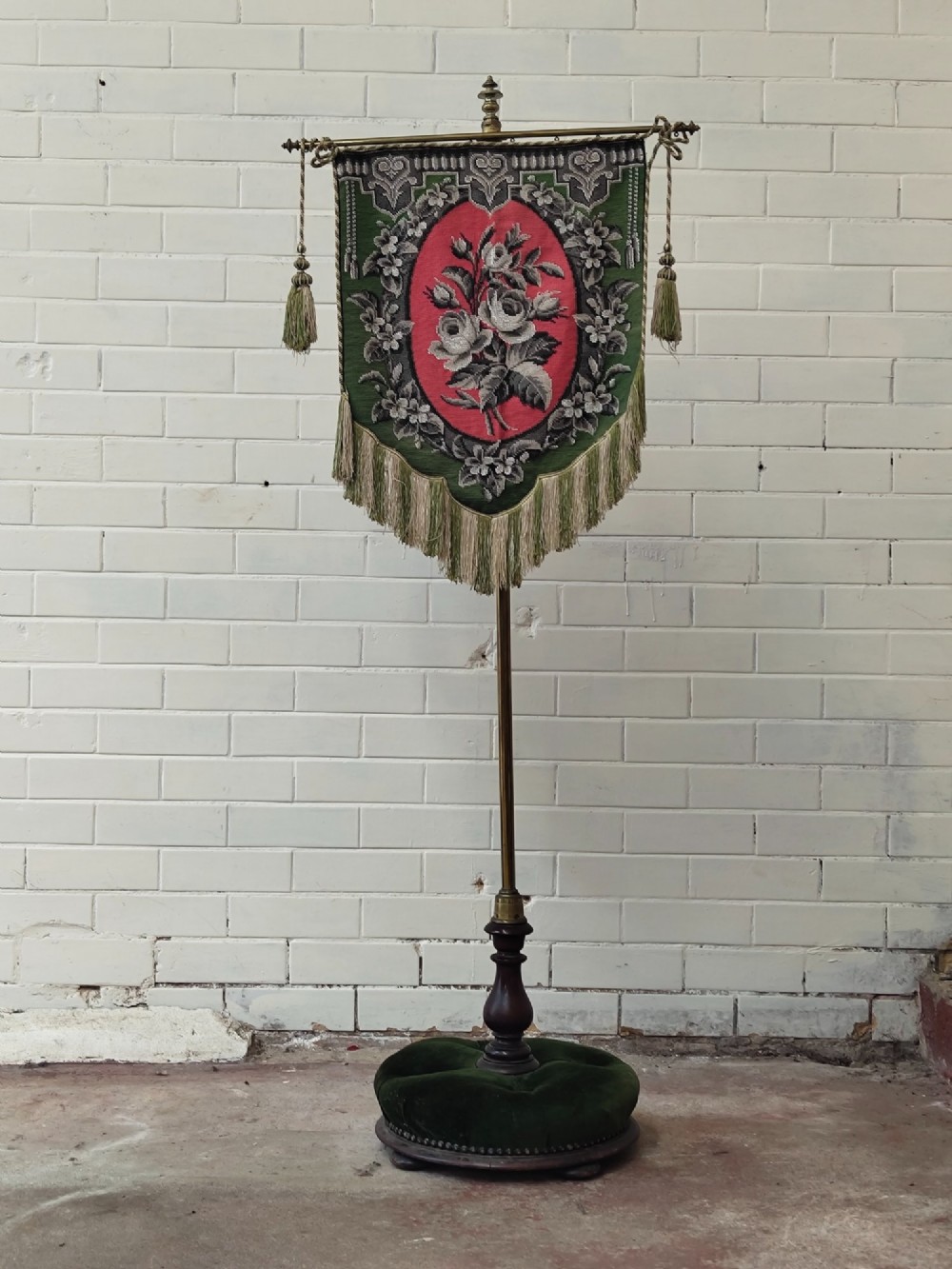 An antique Victorian brass pole screen featuring a green embroidered floral tapestry banner, fringed border, tassels, and a dark green velvet base.