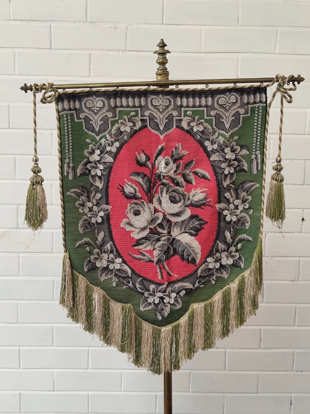 An antique Victorian brass pole screen featuring a green embroidered floral tapestry banner, fringed border, tassels, and a dark green velvet base. View VI