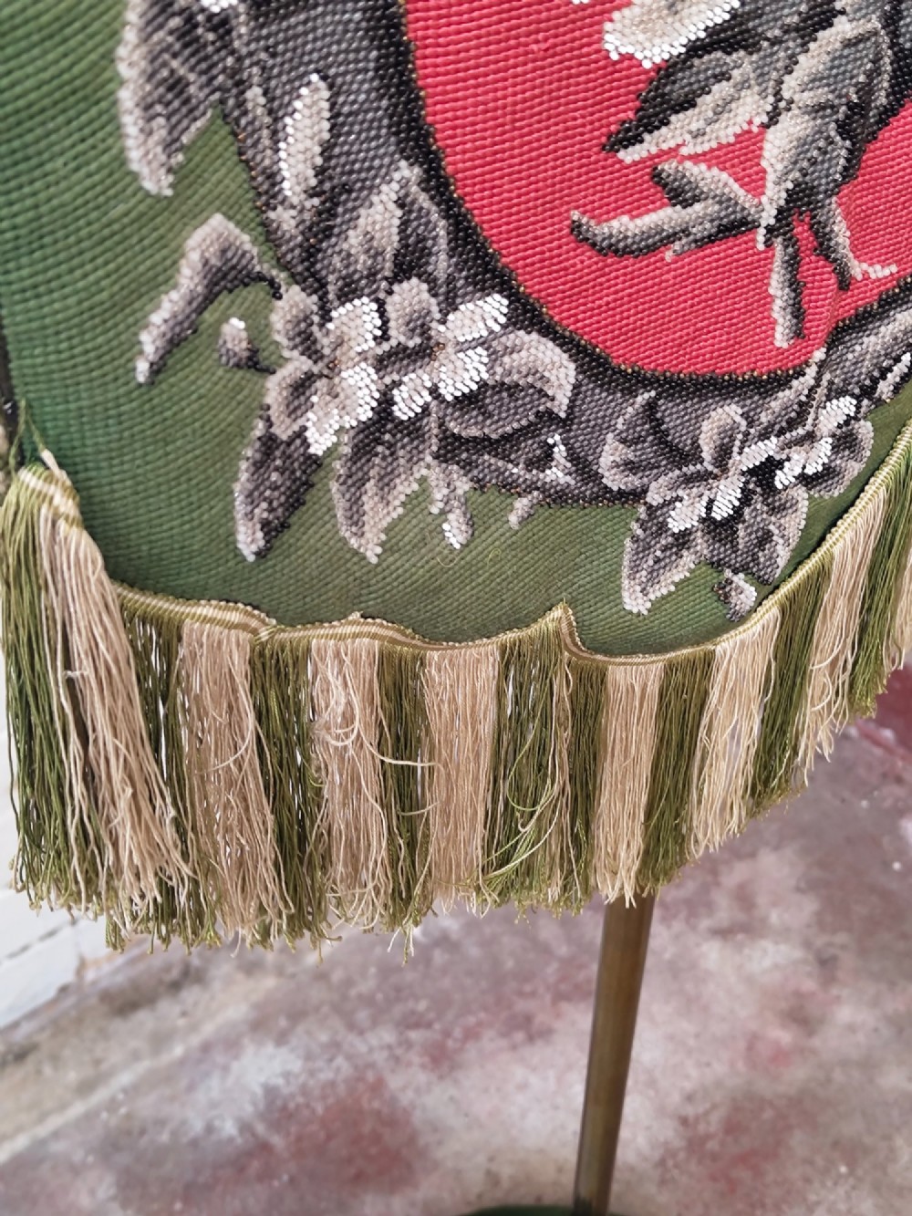 An antique Victorian brass pole screen featuring a green embroidered floral tapestry banner, fringed border, tassels, and a dark green velvet base. View VII