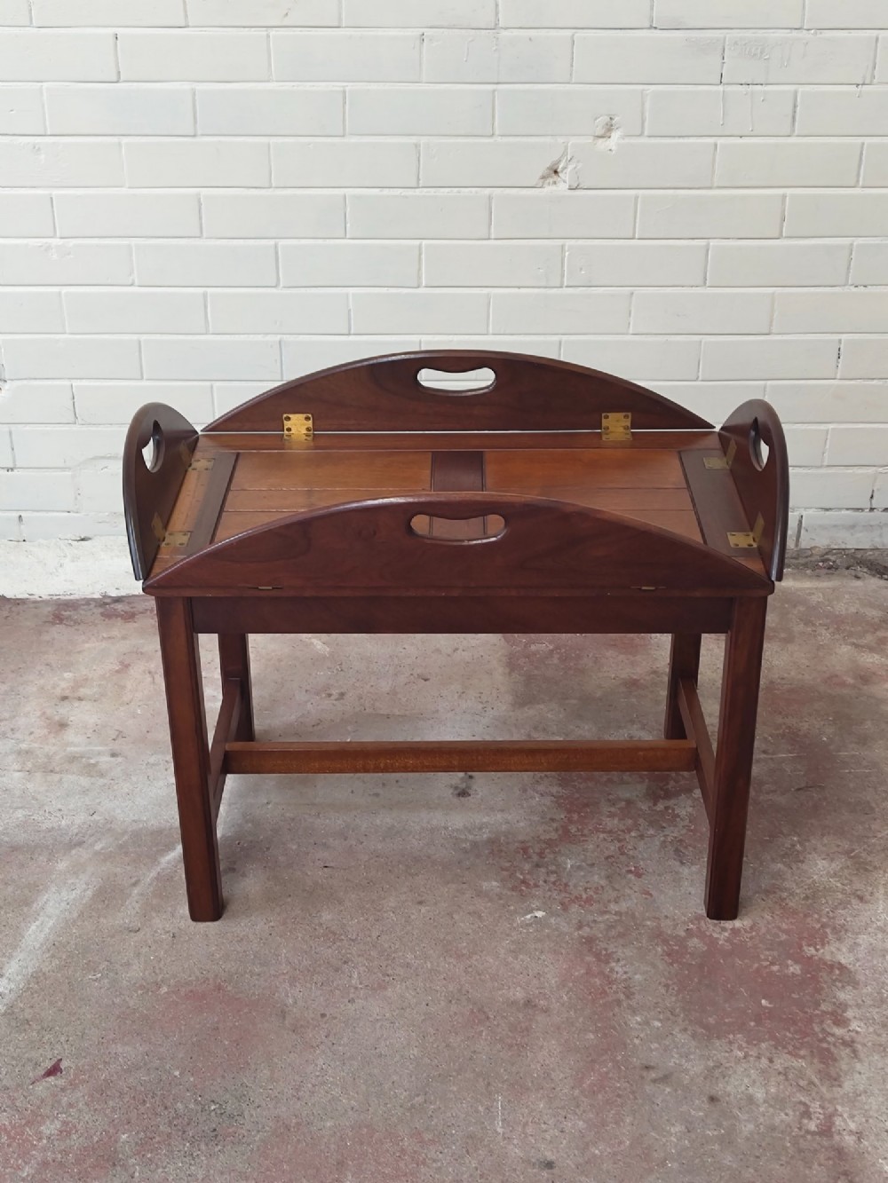 An antique mahogany butlers tray with hinged folding sides and brass fittings resting on a rectangular wooden stand. View II