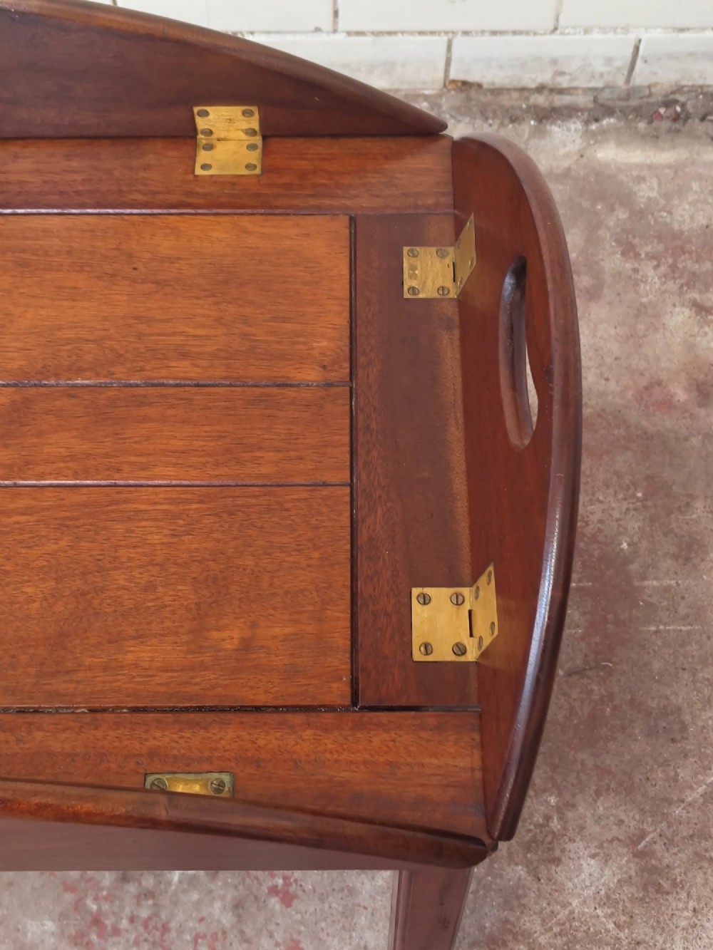 An antique mahogany butlers tray with hinged folding sides and brass fittings resting on a rectangular wooden stand. View V