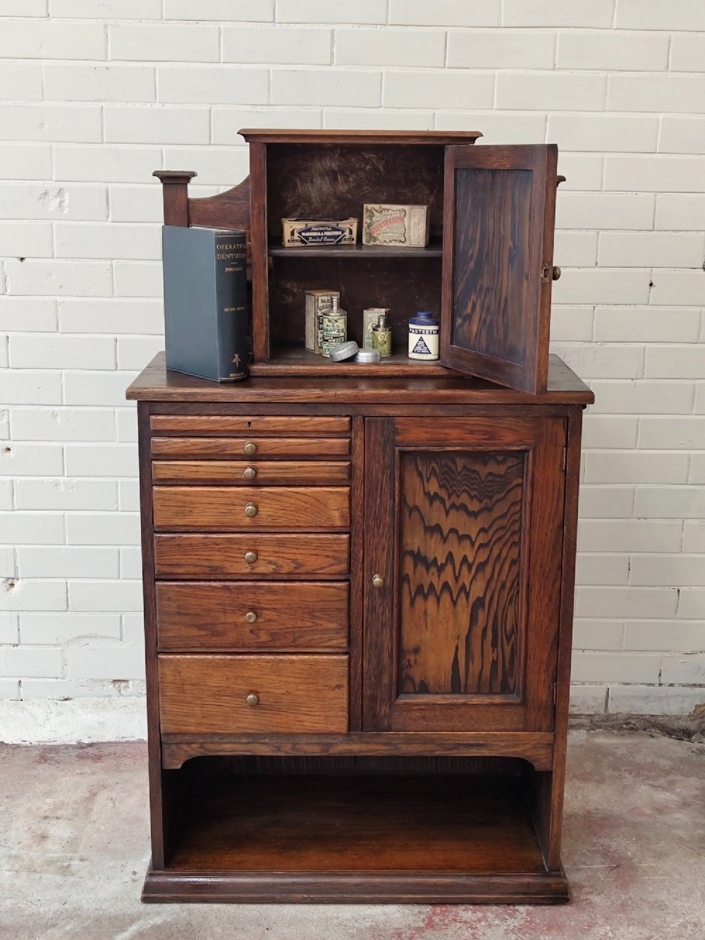 An Edwardian solid oak dentist cabinet featuring multiple shallow drawers, paneled cupboard doors, brass turned knobs, and an open lower shelf.