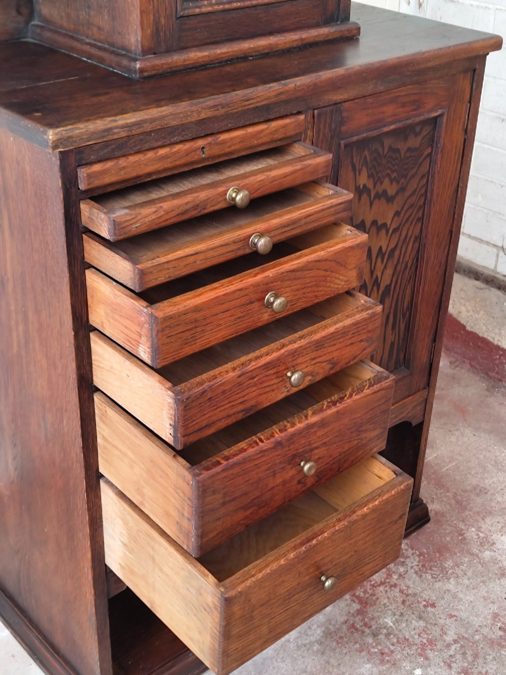 An Edwardian solid oak dentist cabinet featuring multiple shallow drawers, paneled cupboard doors, brass turned knobs, and an open lower shelf. View IX