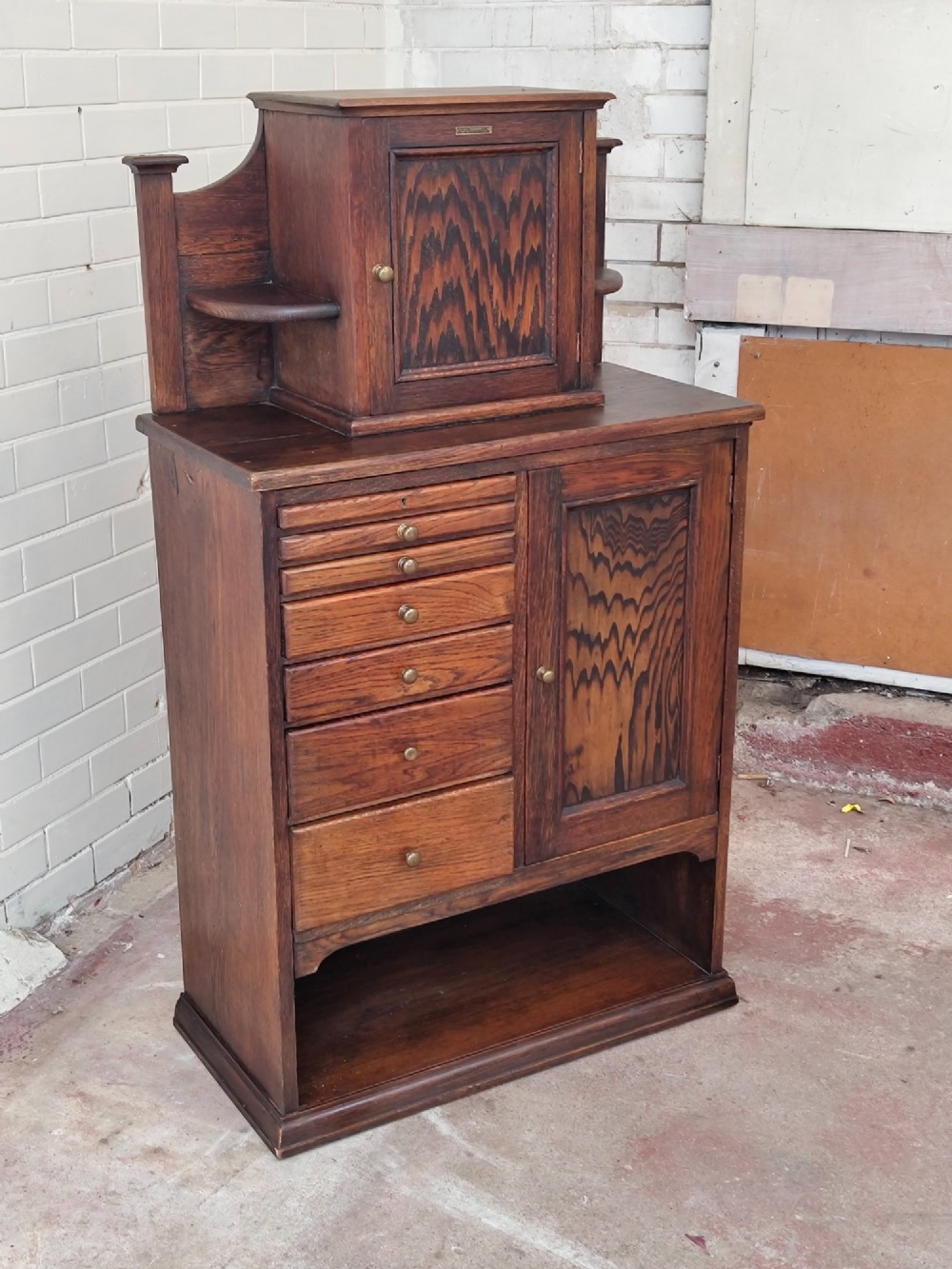 An Edwardian solid oak dentist cabinet featuring multiple shallow drawers, paneled cupboard doors, brass turned knobs, and an open lower shelf. View XI