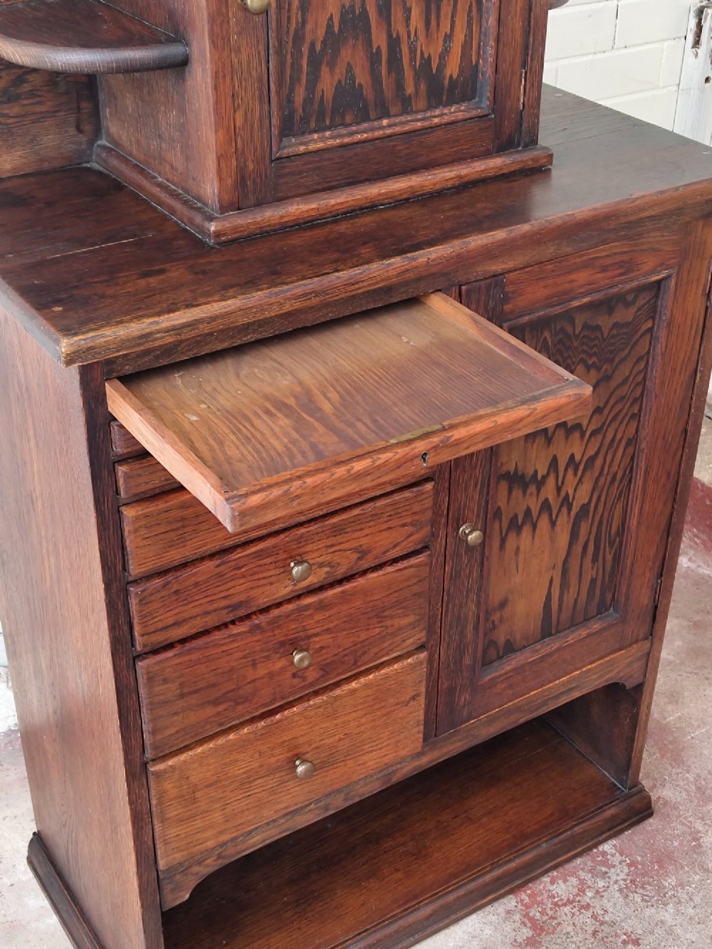 An Edwardian solid oak dentist cabinet featuring multiple shallow drawers, paneled cupboard doors, brass turned knobs, and an open lower shelf. View XV