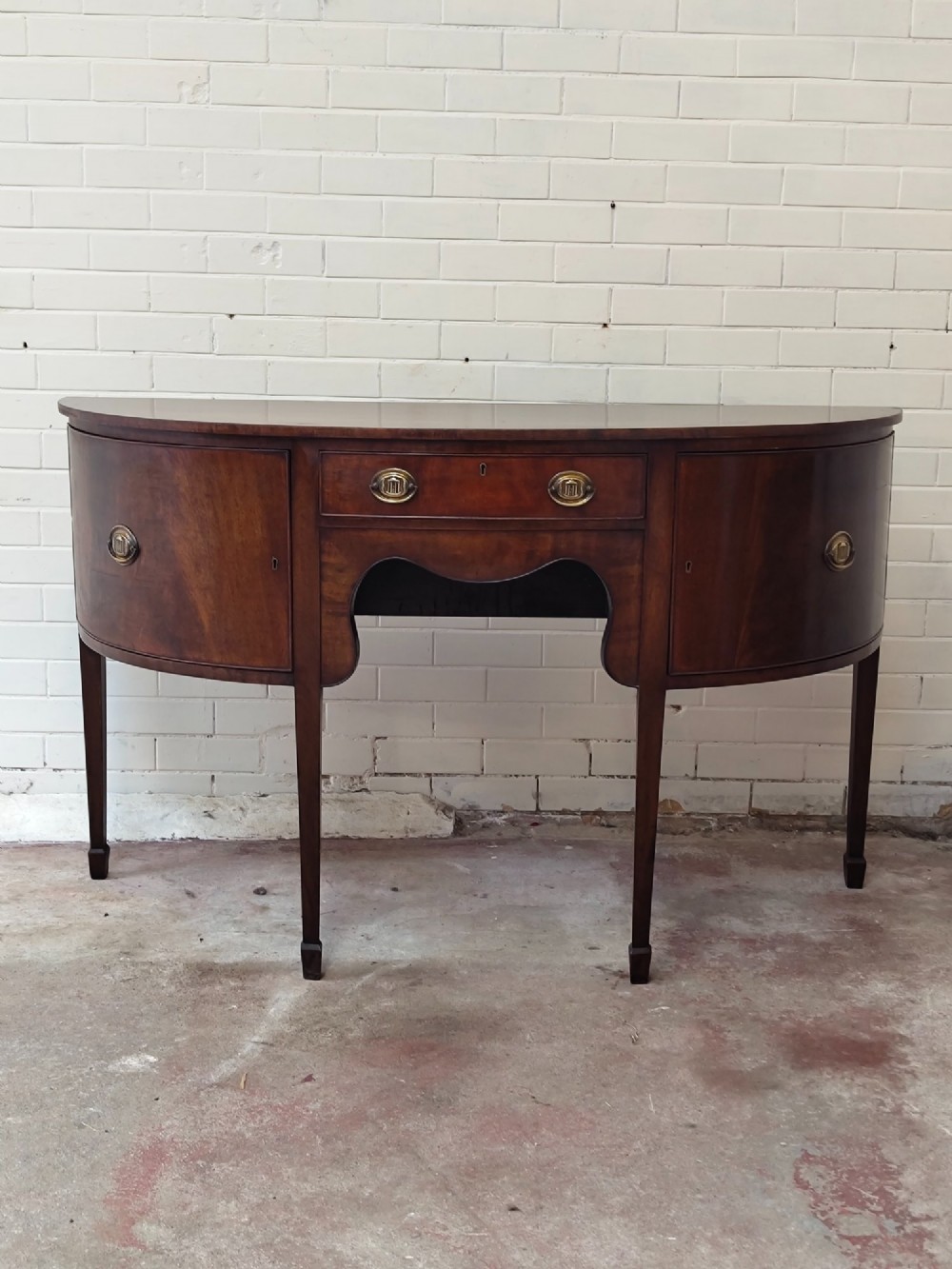 An antique Georgian mahogany demi-lune sideboard featuring a curved front, central drawer, flanking cupboard doors, brass ring handles, and tapered square legs.