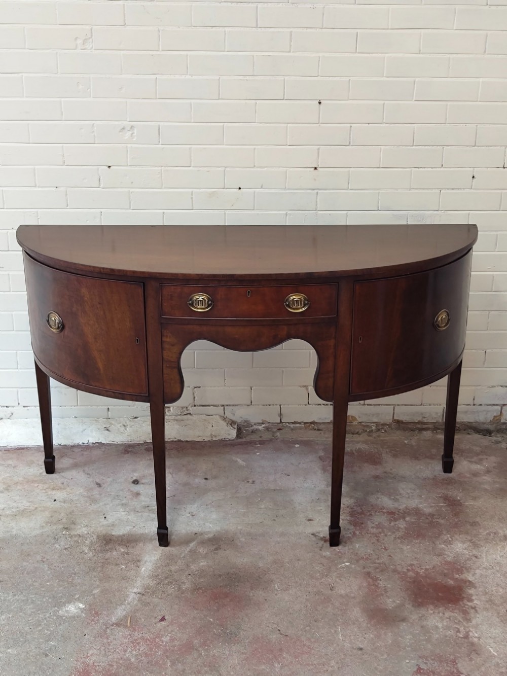 An antique Georgian mahogany demi-lune sideboard featuring a curved front, central drawer, flanking cupboard doors, brass ring handles, and tapered square legs. View II