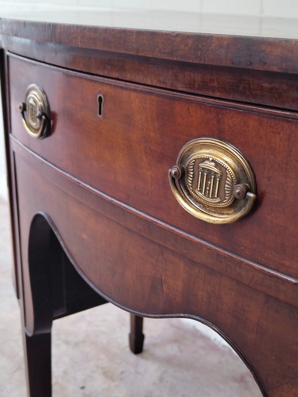 An antique Georgian mahogany demi-lune sideboard featuring a curved front, central drawer, flanking cupboard doors, brass ring handles, and tapered square legs. View IV