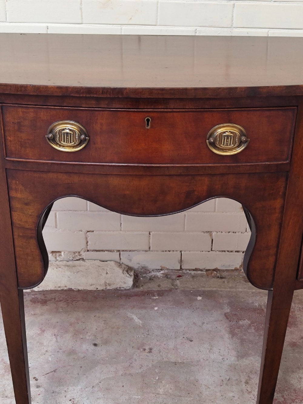 An antique Georgian mahogany demi-lune sideboard featuring a curved front, central drawer, flanking cupboard doors, brass ring handles, and tapered square legs. View V