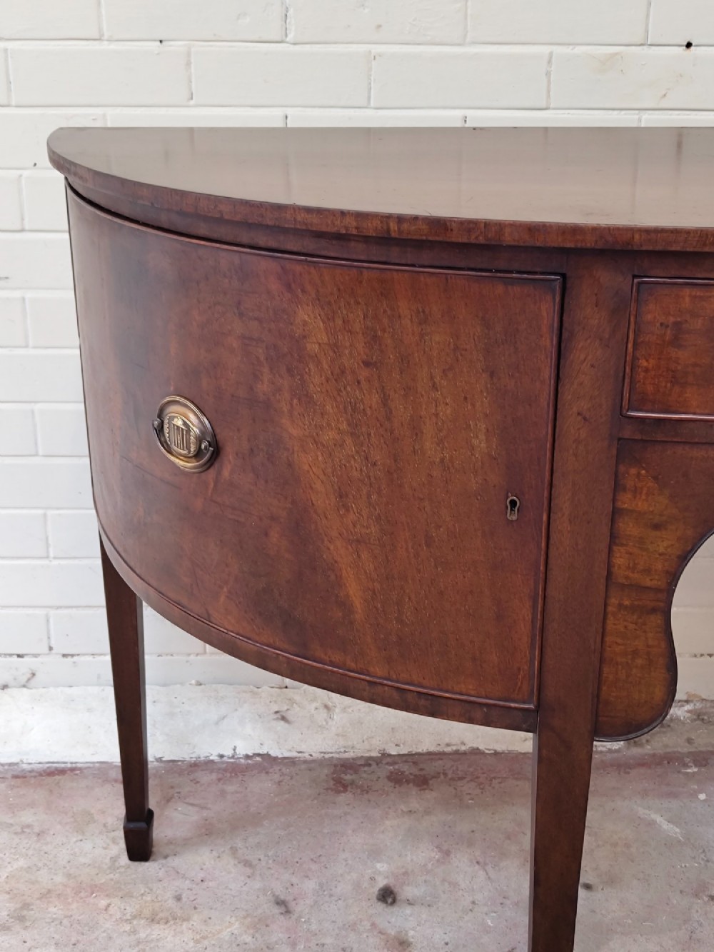 An antique Georgian mahogany demi-lune sideboard featuring a curved front, central drawer, flanking cupboard doors, brass ring handles, and tapered square legs. View VI