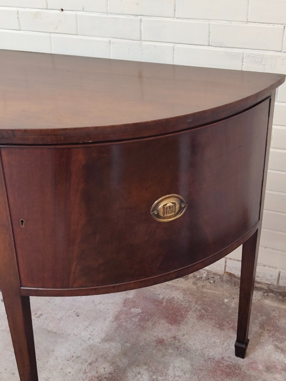 An antique Georgian mahogany demi-lune sideboard featuring a curved front, central drawer, flanking cupboard doors, brass ring handles, and tapered square legs. View VII