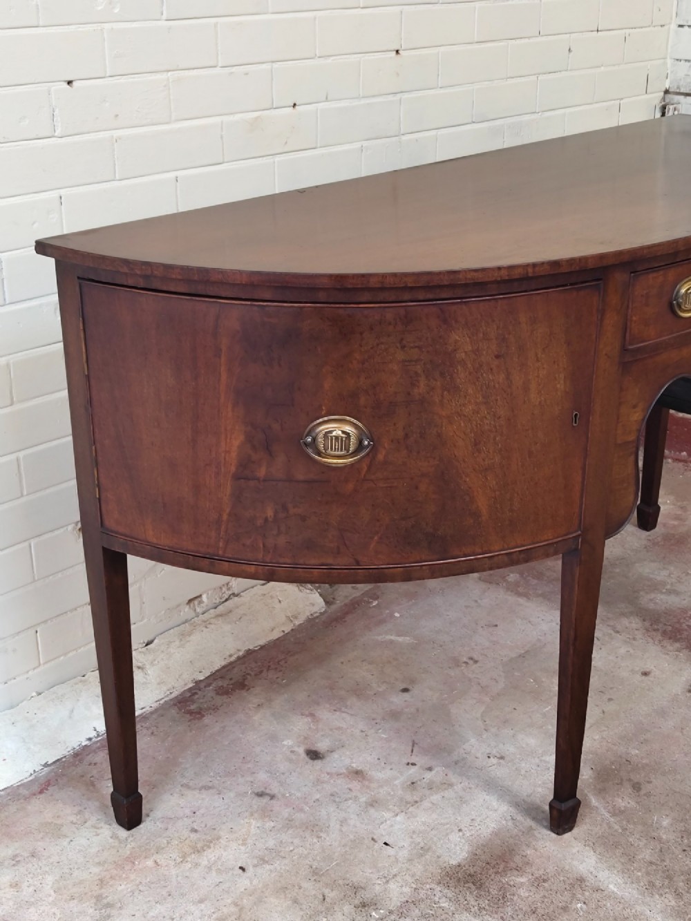 An antique Georgian mahogany demi-lune sideboard featuring a curved front, central drawer, flanking cupboard doors, brass ring handles, and tapered square legs. View VIII