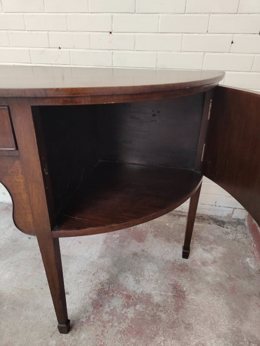 An antique Georgian mahogany demi-lune sideboard featuring a curved front, central drawer, flanking cupboard doors, brass ring handles, and tapered square legs. View IX
