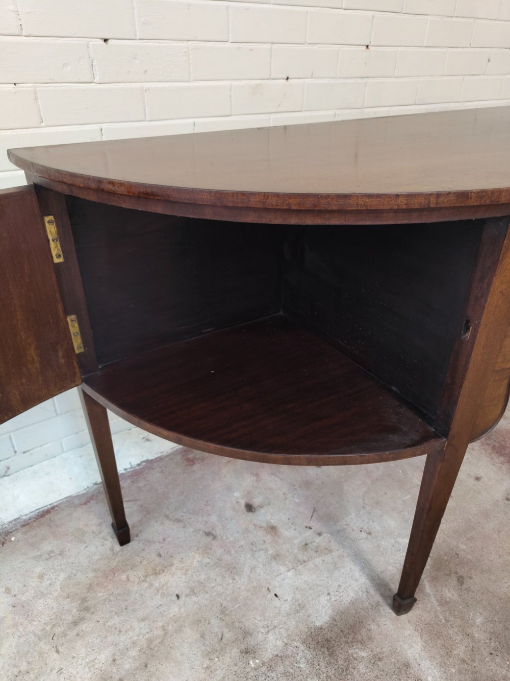 An antique Georgian mahogany demi-lune sideboard featuring a curved front, central drawer, flanking cupboard doors, brass ring handles, and tapered square legs. View X