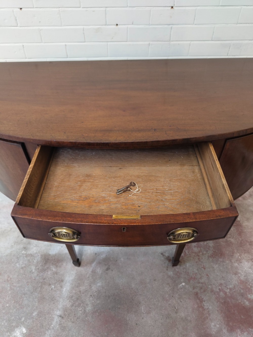 An antique Georgian mahogany demi-lune sideboard featuring a curved front, central drawer, flanking cupboard doors, brass ring handles, and tapered square legs. View XI