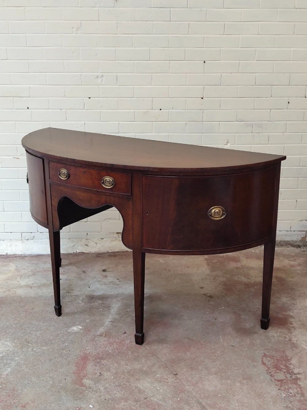 An antique Georgian mahogany demi-lune sideboard featuring a curved front, central drawer, flanking cupboard doors, brass ring handles, and tapered square legs. View XII