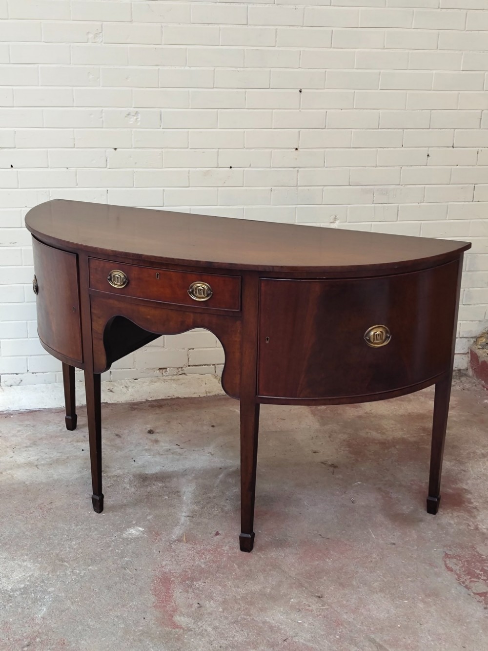 An antique Georgian mahogany demi-lune sideboard featuring a curved front, central drawer, flanking cupboard doors, brass ring handles, and tapered square legs. View XIII
