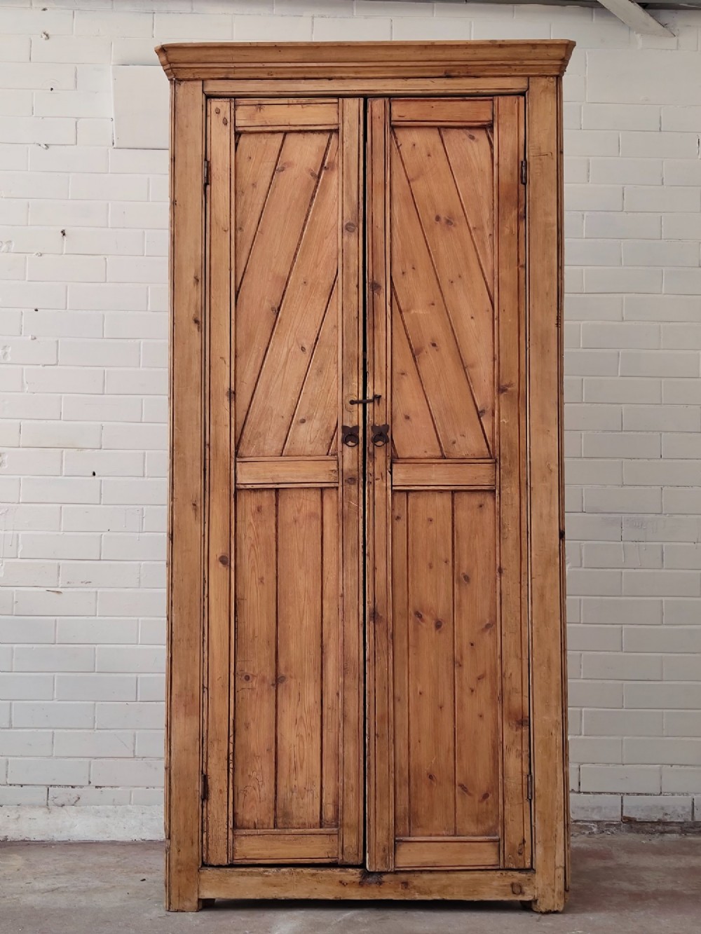 A tall antique pitch pine corner wardrobe featuring double doors with diagonal timber panels and a moulded top cornice.