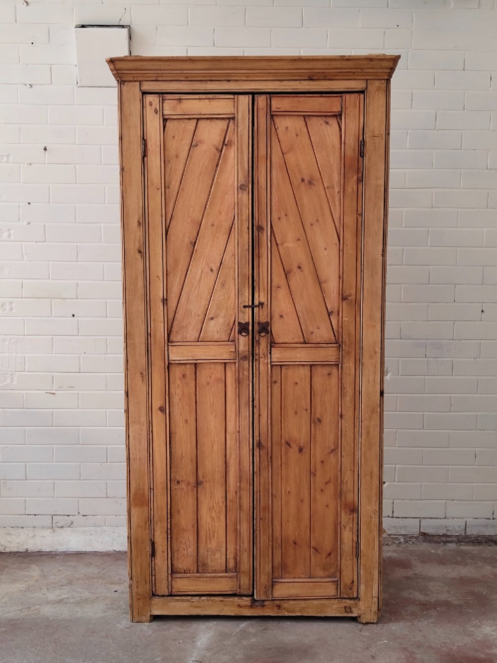 A tall antique pitch pine corner wardrobe featuring double doors with diagonal timber panels and a moulded top cornice. View II