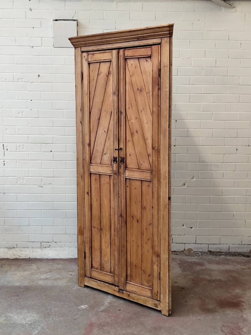 A tall antique pitch pine corner wardrobe featuring double doors with diagonal timber panels and a moulded top cornice. View XV