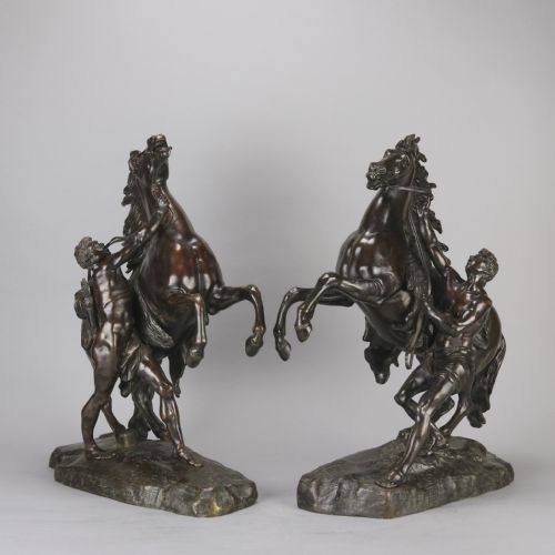 fig 1. A pair of dark brown bronze sculptures depicting rearing horses restrained by standing male figures, set on naturalistic rock bases.