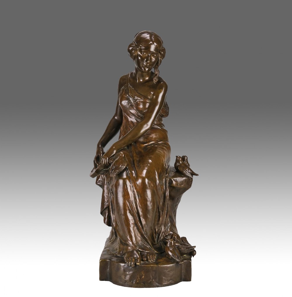 A dark brown patinated bronze sculpture depicting a seated woman in classical draped attire interacting with small birds, set on an integral bronze base.