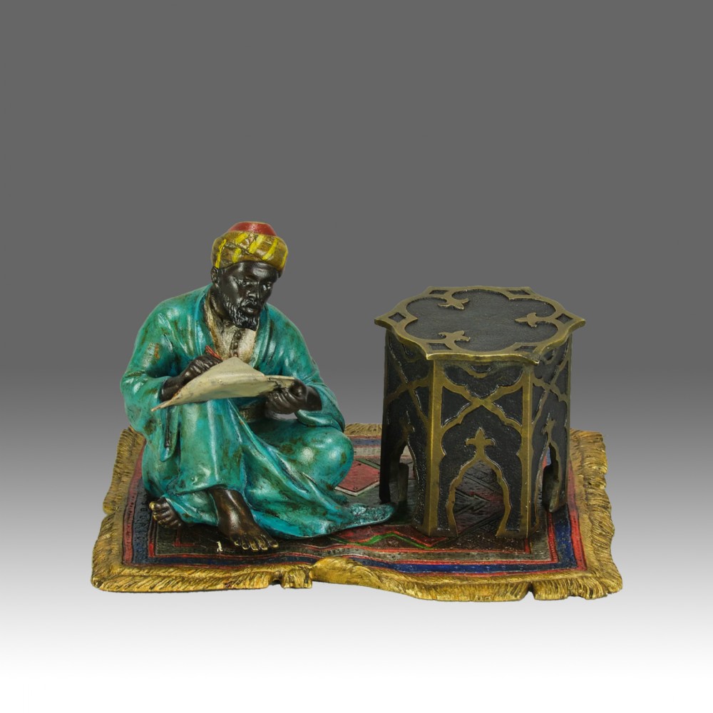 A Viennese cold-painted bronze sculpture depicting a seated scholar in a turquoise robe writing beside an octagonal Moorish table on a patterned carpet.