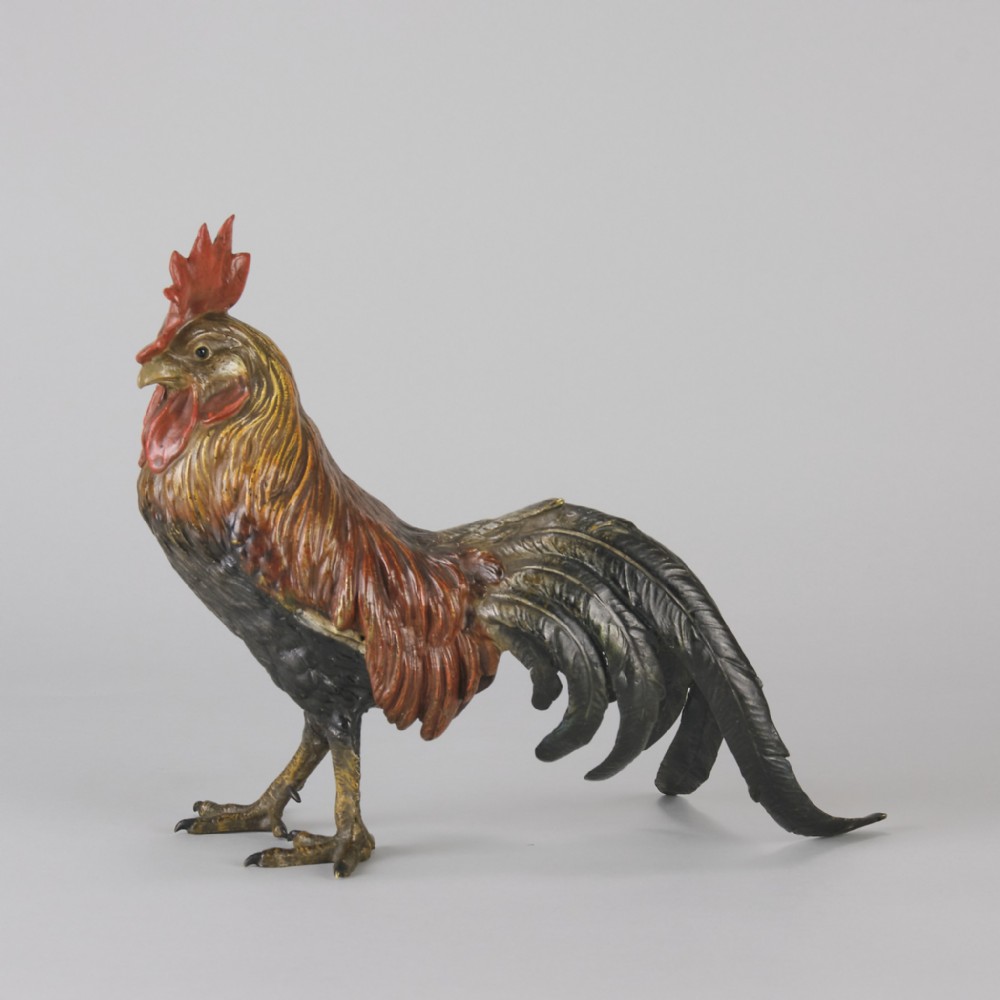 A cold-painted bronze figurine depicting a standing cockerel with a red comb and textured feathers in brown, ochre, and dark grey shades.