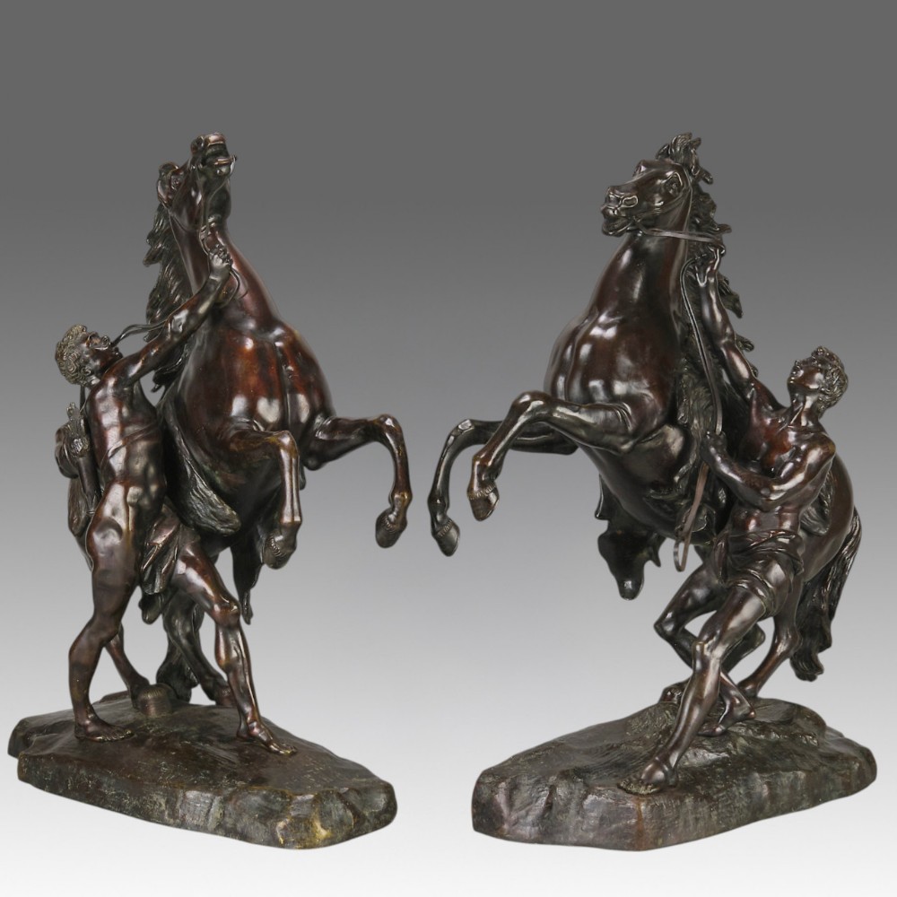A pair of dark brown bronze sculptures depicting rearing horses restrained by standing male figures, set on naturalistic rock bases.