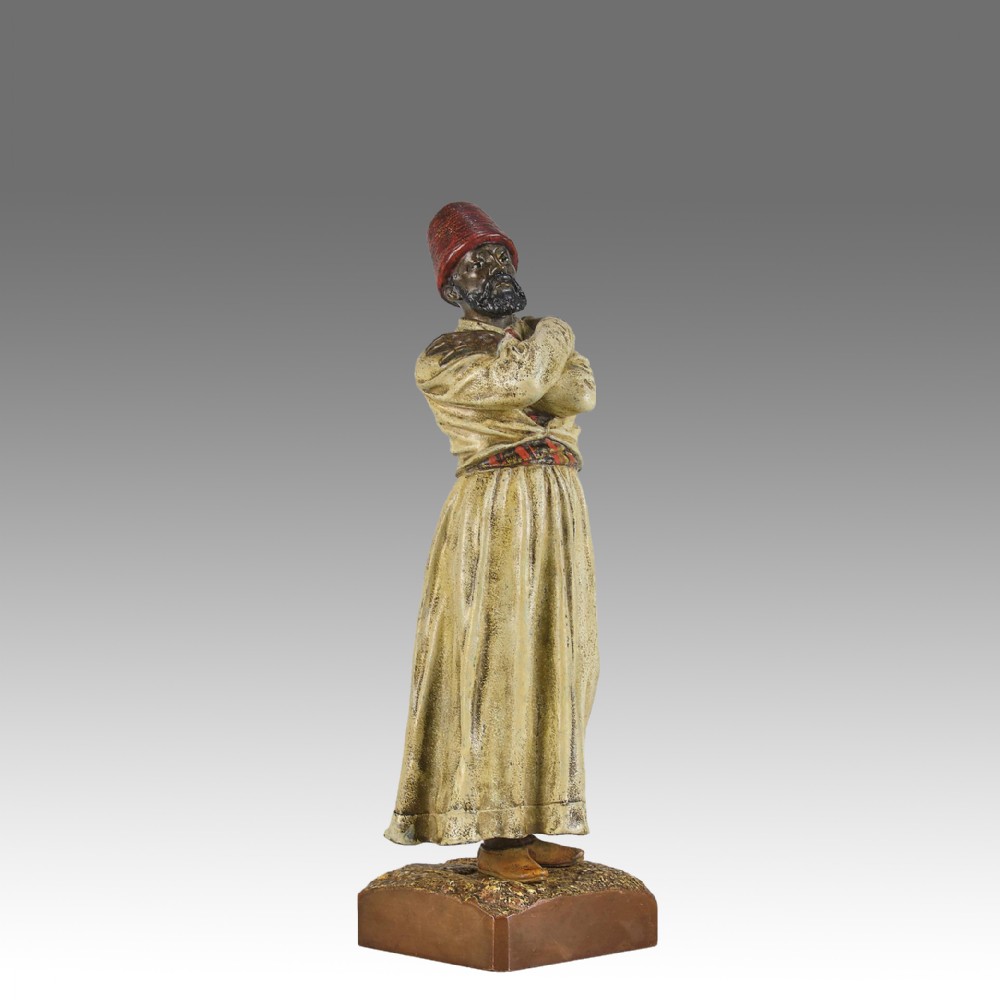 An early twentieth-century cold-painted bronze figure depicting a bearded man in a cream robe and red fez with arms crossed on a rectangular base.