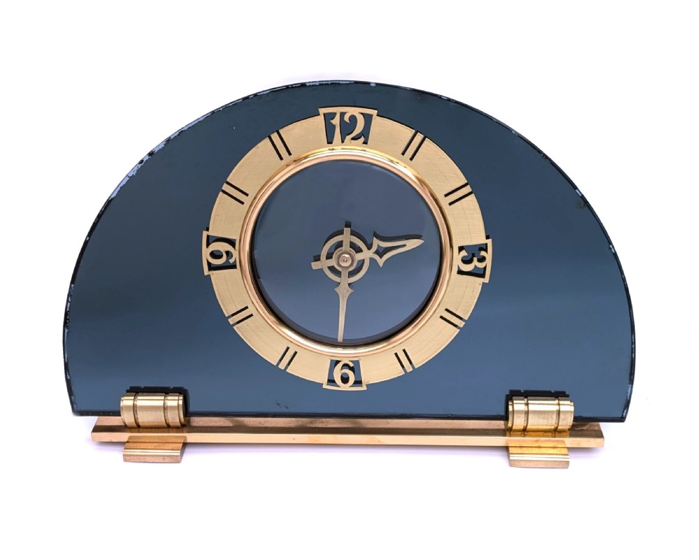 An Art Deco mantel clock featuring a semi-circular blue glass body, a circular brass dial with stylized numerals, and a blue braided flex. View II